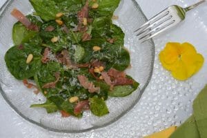 A plate of Warm Spinach Basil Salad with strips of pancetta and toasted pine nuts plus a garnish of a yellow pansy.