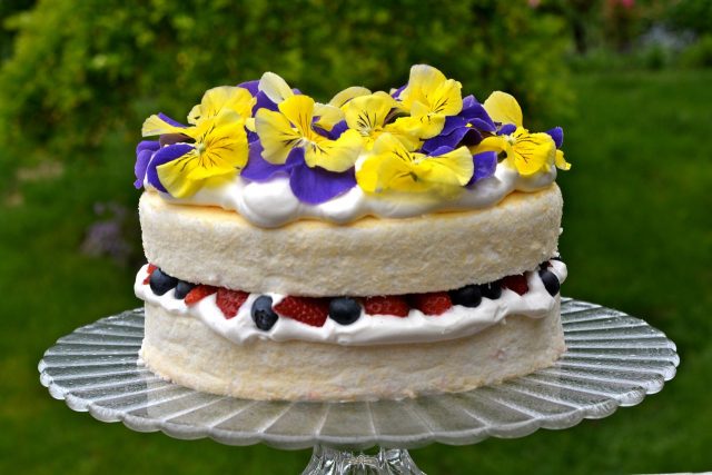 A pedestal tray with a gluten free Angel Food Cake filled with whipped cream and fresh berries, garnished with pansies.