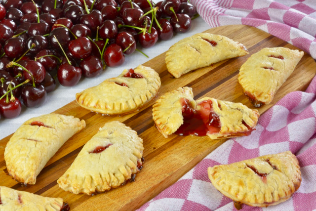 A wooden board covered in Cherry Hand Pies surrounded by an red and white checked tea towel.