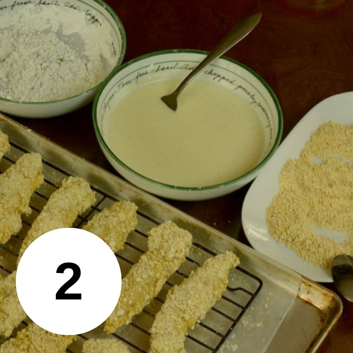 Three bowls with cornstarch, egg wash and a cornflake crumb mixture. Each bowl holds a chicken strip and a metal baking tray with wire rack holds 4 completed pieces of breaded chicken.