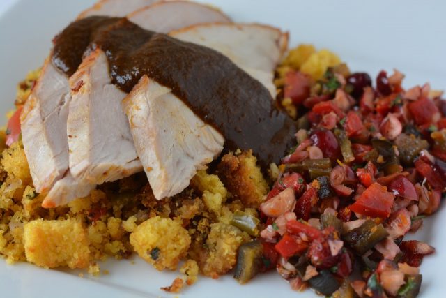 A plate of turkey slices on top of gluten free cornbread sausage stuffing topped with ancho chile gravy and cranberry salsa on the side.