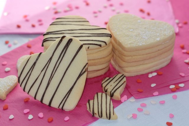 A stack of heart shaped gluten free sugar cookies drizzled with chocolate sitting on pretty pink paper surrounded by tiny heart shaped sprinkles.