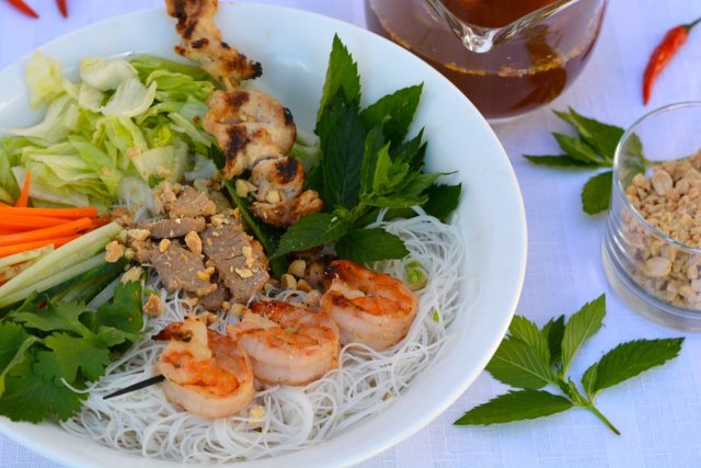 A white bowl filled with rice noodles, grilled shrimp, chicken and pork, shredded lettuce and fresh herbs with a dish of nuoc cham dipping sauce beside it.