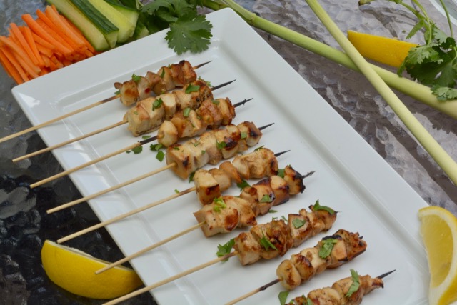 A platter with skewers of Lemongrass Chicken surrounded by lemongrass, carrots, cucumber and lemon wedges.