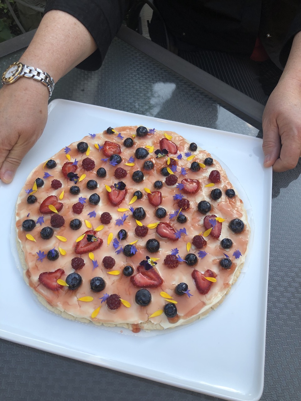 Two hands holding a square white platter holding a glazed fruit pizza that has randomly placed strawberries, raspberries and blueberries. Finished with a sprinkling of blue and yellow. edible flower petals.