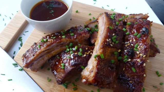 A cutting board with hoisin glazed pork ribs sprinkled with sesame seeds and chopped green onion with extra sauce on the side.