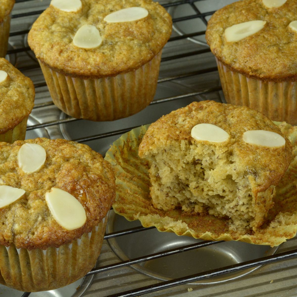 A baking rack with Banana Muffins each topped with 3 pieces of sliced almonds.