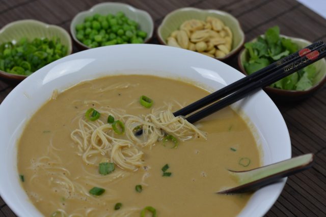 A bowl of Noodle Soup with Peanut Sauce surrounded by optional toppings.