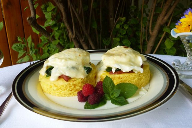 A white plate with gluten free cornbread rounds topped with ham, spinach, eggs and Mornay sauce, Eggs Florentine.