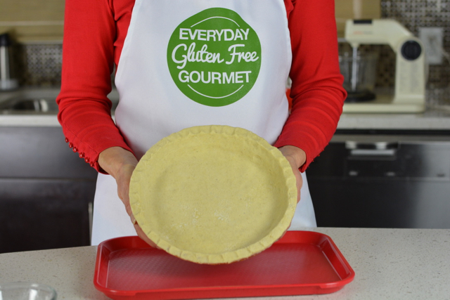 Cinde wearing her Everyday Gluten Free Gourmet apron holding a raw pie crust in a pie plate.