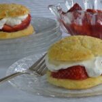 Strawberry Shortcake on a plate filled with strawberry sauce and whipped cream, with more in the background.