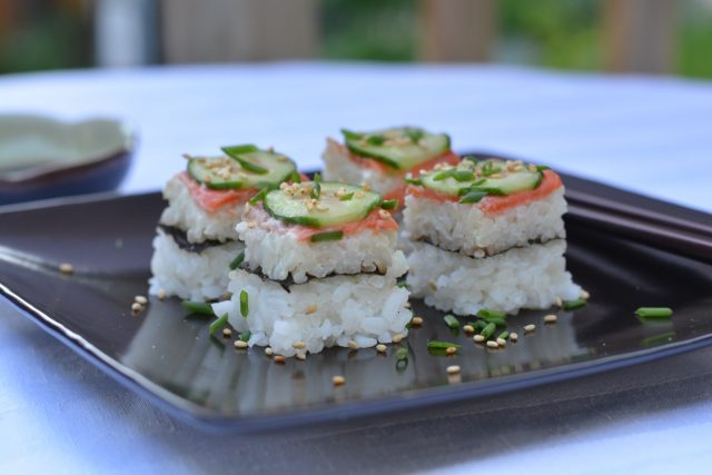 A black tray of smoked salmon sushi pizza cut in squares and sprinkled with snipped chives and sesame seeds, accompanied by gluten free soy sauce.