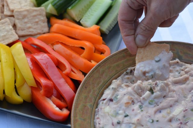 A cracker being dipped into warm Cranberry Dip surrounded by sliced raw veggies.