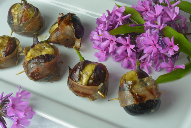 Grilled Figs Stuffed with Blue Cheese and wrapped with prosciutto on a white platter surrounded by pink flowers