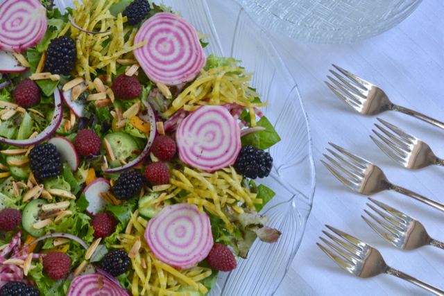 A glass bowl with Garden Harvest Jewelled Salad containing sliced candy cane beets, grated golden beets, raspberries, blackberries and a variety of lettuce.