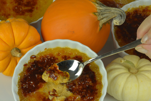 A dish of Pumpkin Creme Brulee with the first bite on the spoon surrounded by orange and white mini pumpkins.