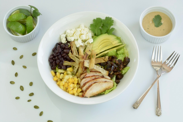 A white bowl filled with Santa Fe Salad; blackened chicken slices, avocado, corn, beans, feta cheese and dates.