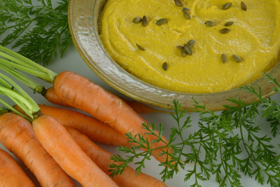 Roasted Carrot Hummus in a round dish sprinkled with toasted pumpkin seeds and surrounded by garden carrots.