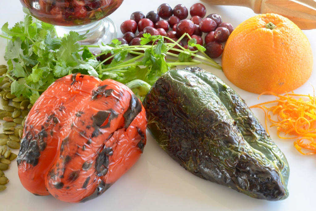 A roasted red pepper and a roasted poblano surround by cilantro, cranberries and an orange.