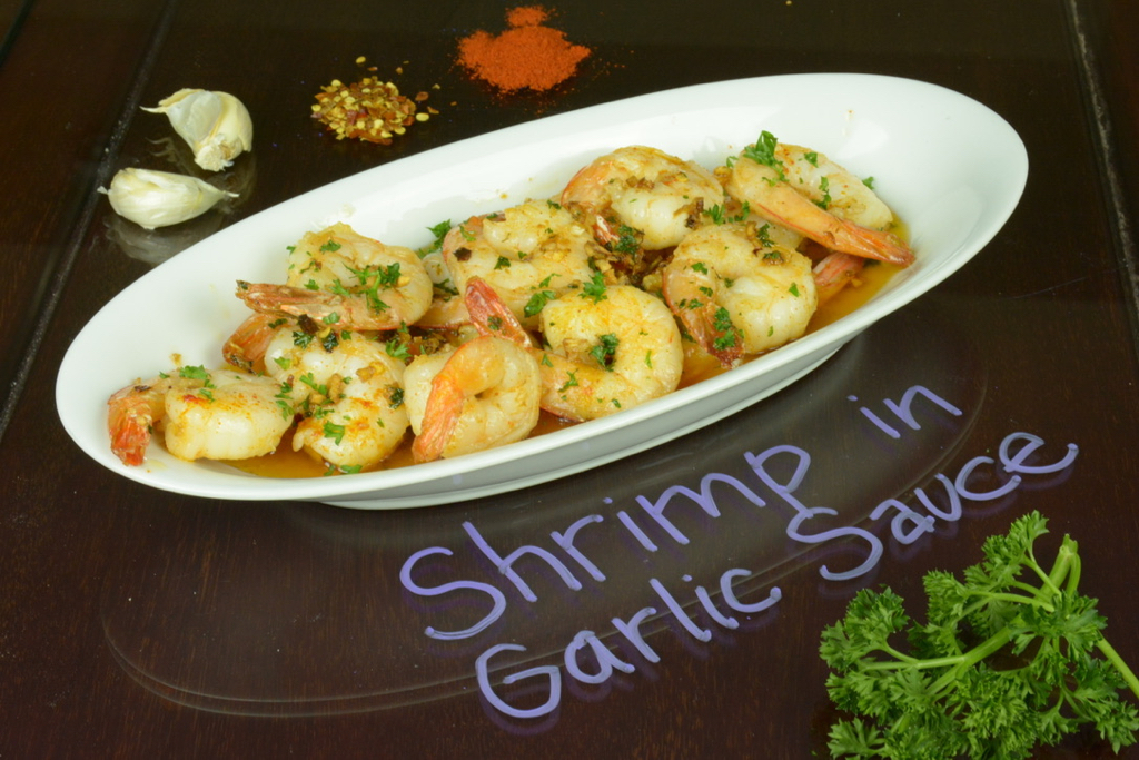 White plate with shrimp in garlic sauce sprinkled with parsley, in the background is garlic, chile flakes and paprika.