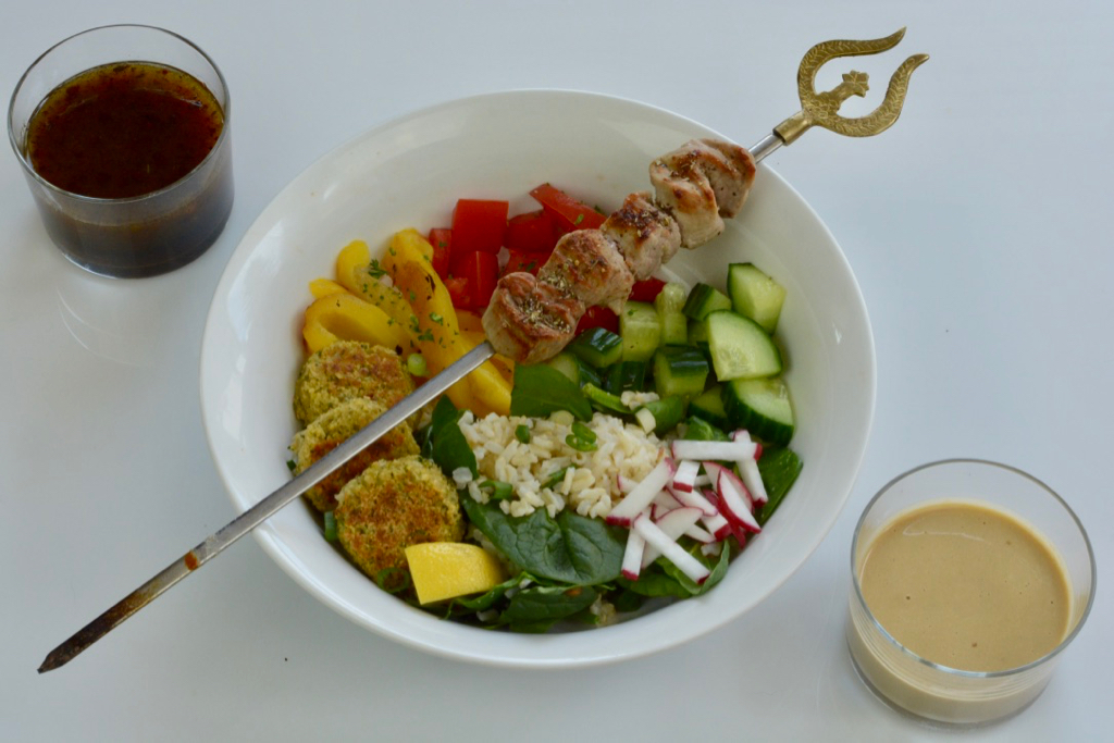 A white bowl filled with Middle Eastern delights; falafel balls, chopped vegetables and rice topped with a pork kebab with tahini sauce on the side.