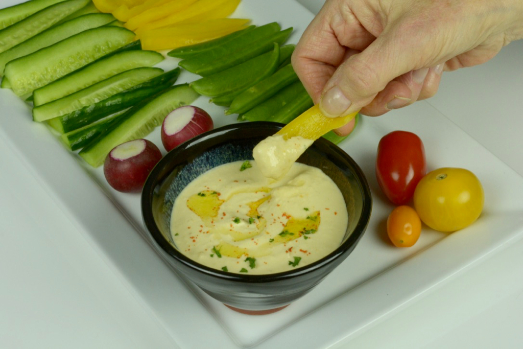 A bowl with Lemony Hummus, surrounded by raw veggies, and a hand dipping a yellow pepper into the hummus.