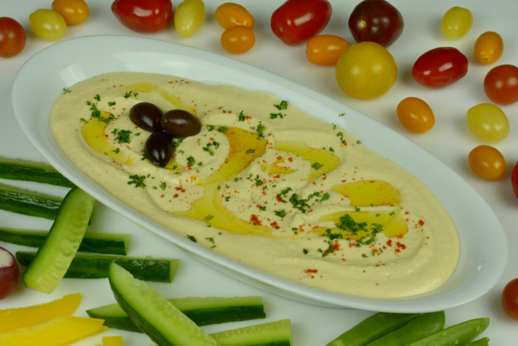 A white, oval shaped dish filled with lemony hummus surrounded by cherry tomatoes and cucumber slices.