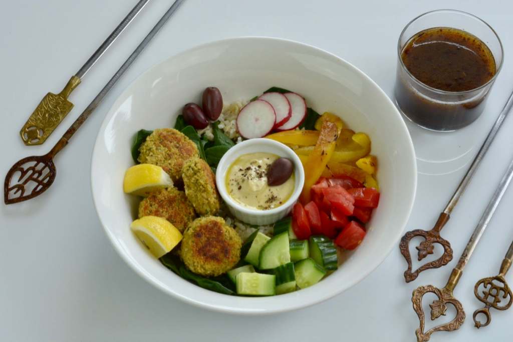 A white bowl with rice, falafel balls, hummus and chopped vegetables with pomegranate dressing on the side.