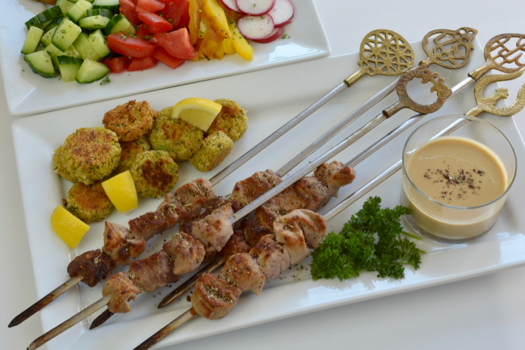 A white platter of Pork Kebabs with Za'atar spices plus falafel balls and cut up vegetables with a dish of tahini dressing..