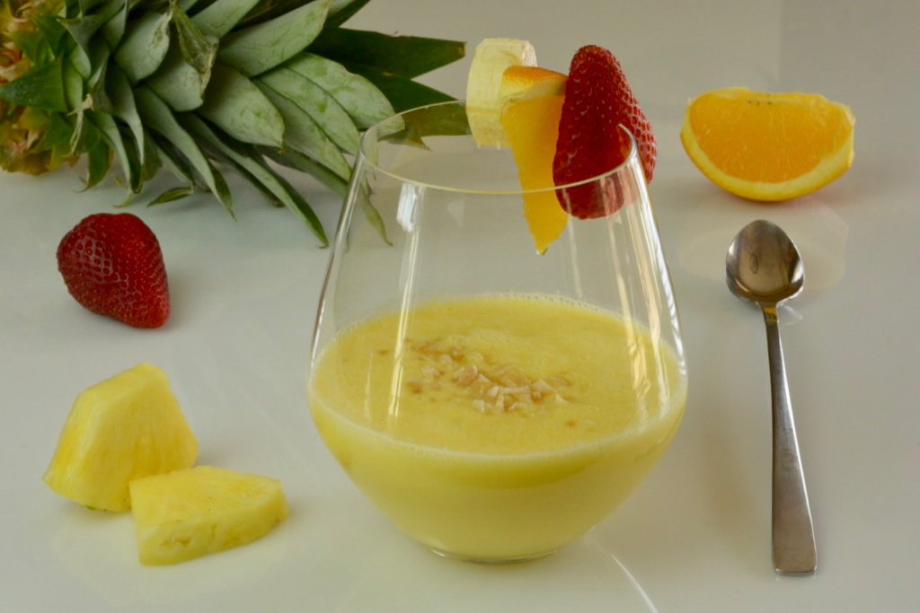 A stemless wine glass with a Tropical Yellow Smoothie sprinkled with toasted coconut and garnished with banana, orange and strawberry.