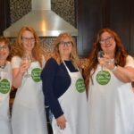 4 ladies wearing aprons in the Everyday Gluten Free Gourmet kitchen raising a glass of wine.