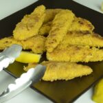 A black plate with breaded chicken strips and lemon wedges ready to serve.
