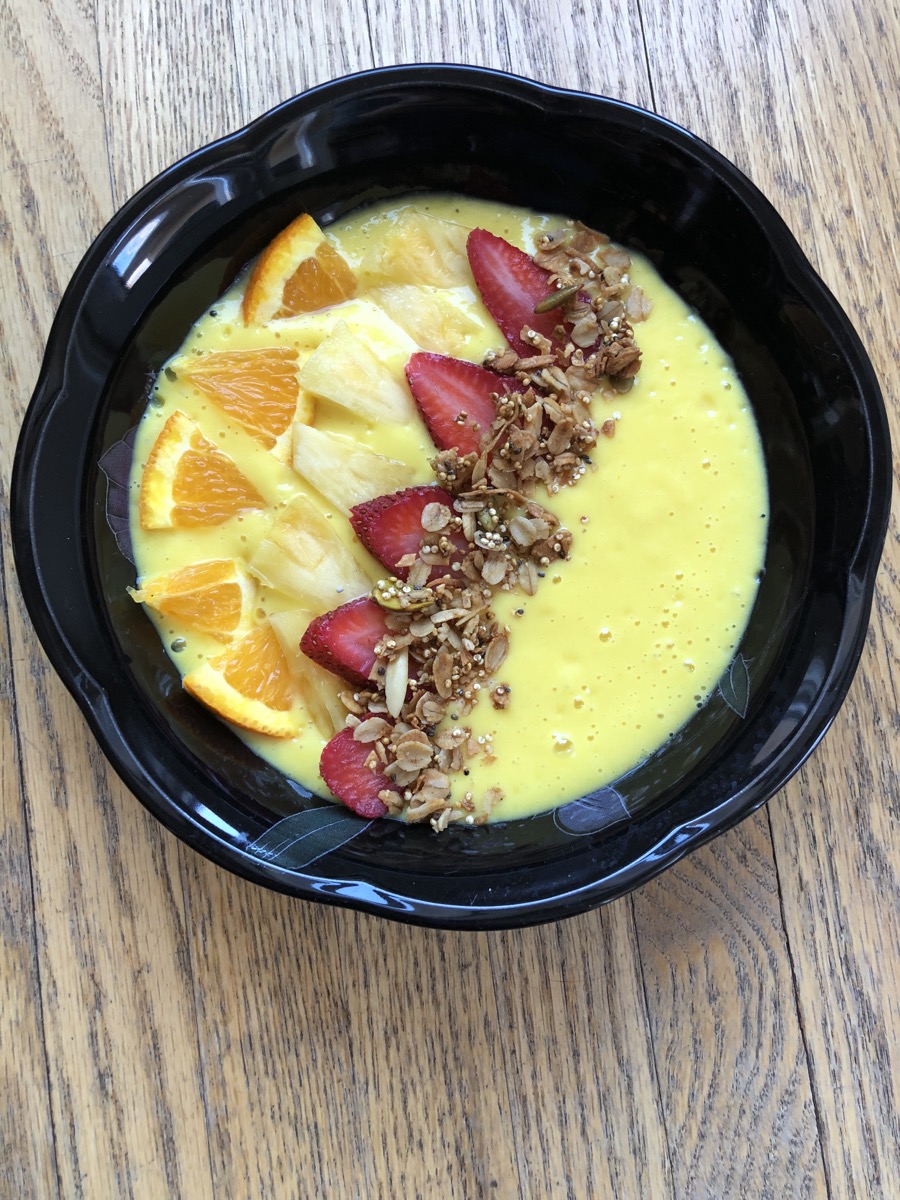 A black bowl with yellow smoothie topped with sliced orange, pineapple and strawberries then sprinkled with granola.