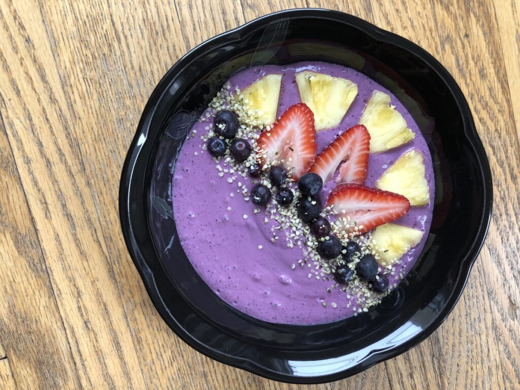 A black bowl filled with an indigo coloured smoothie, topped with sliced pineapple and strawberries, frozen blueberries and a sprinkling of hemp seeds.