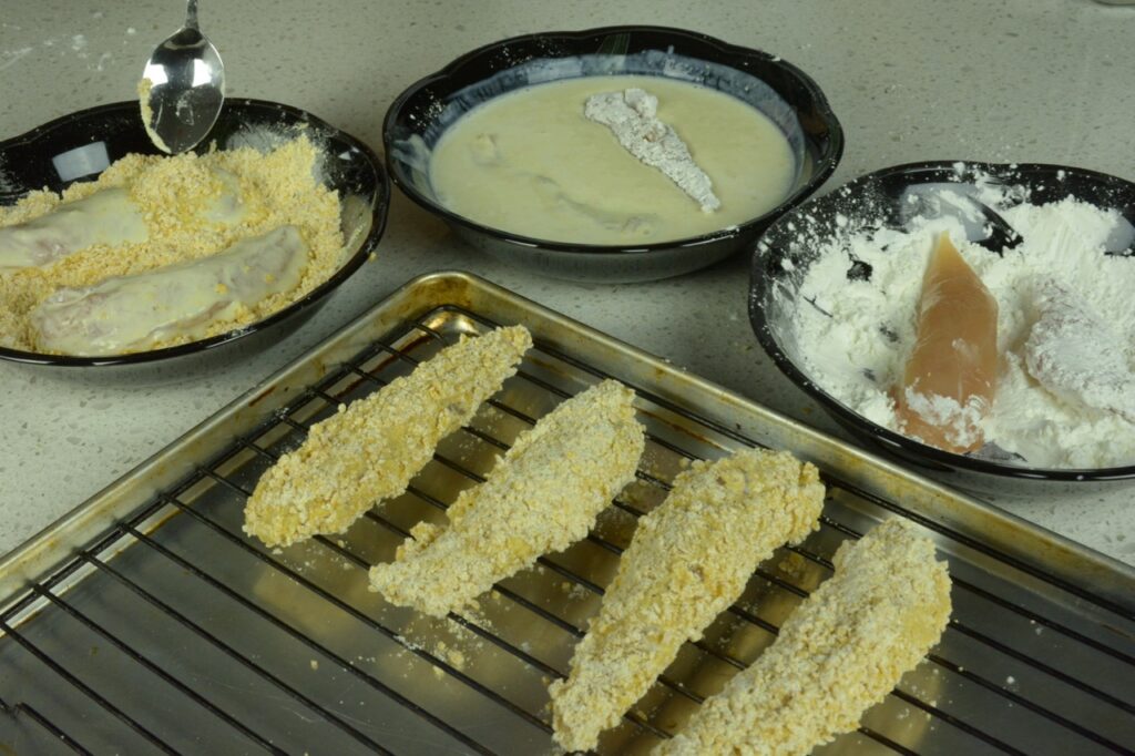 Three black bowls with cornstarch, egg wash and a cornflake crumb mixture. Each bowl holds a chicken strip and a metal baking tray with wire rack holds 4 completed pieces of breaded chicken.