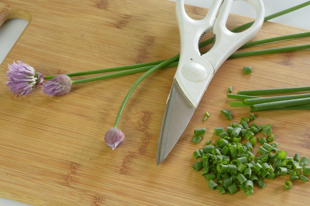 A bamboo cutting board with 3 stems of chives, scissors and a pile of freshly snipped chives.