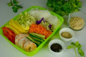 A green container with all the fixings for salad rolls: sliced peppers and cucumber, grated carrots, shrimp halves, cabbage and rice noodles. Beside are dishes with sesame seeds and chopped peanuts.