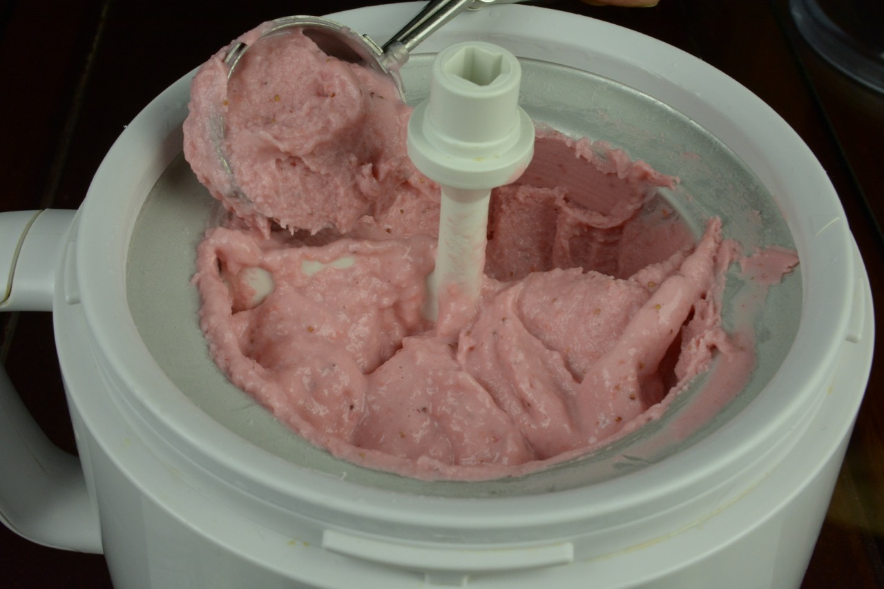 The freezer insert of an ice cream maker filled with soft Strawberry Cheesecake Ice Cream and a scoop taking some out.