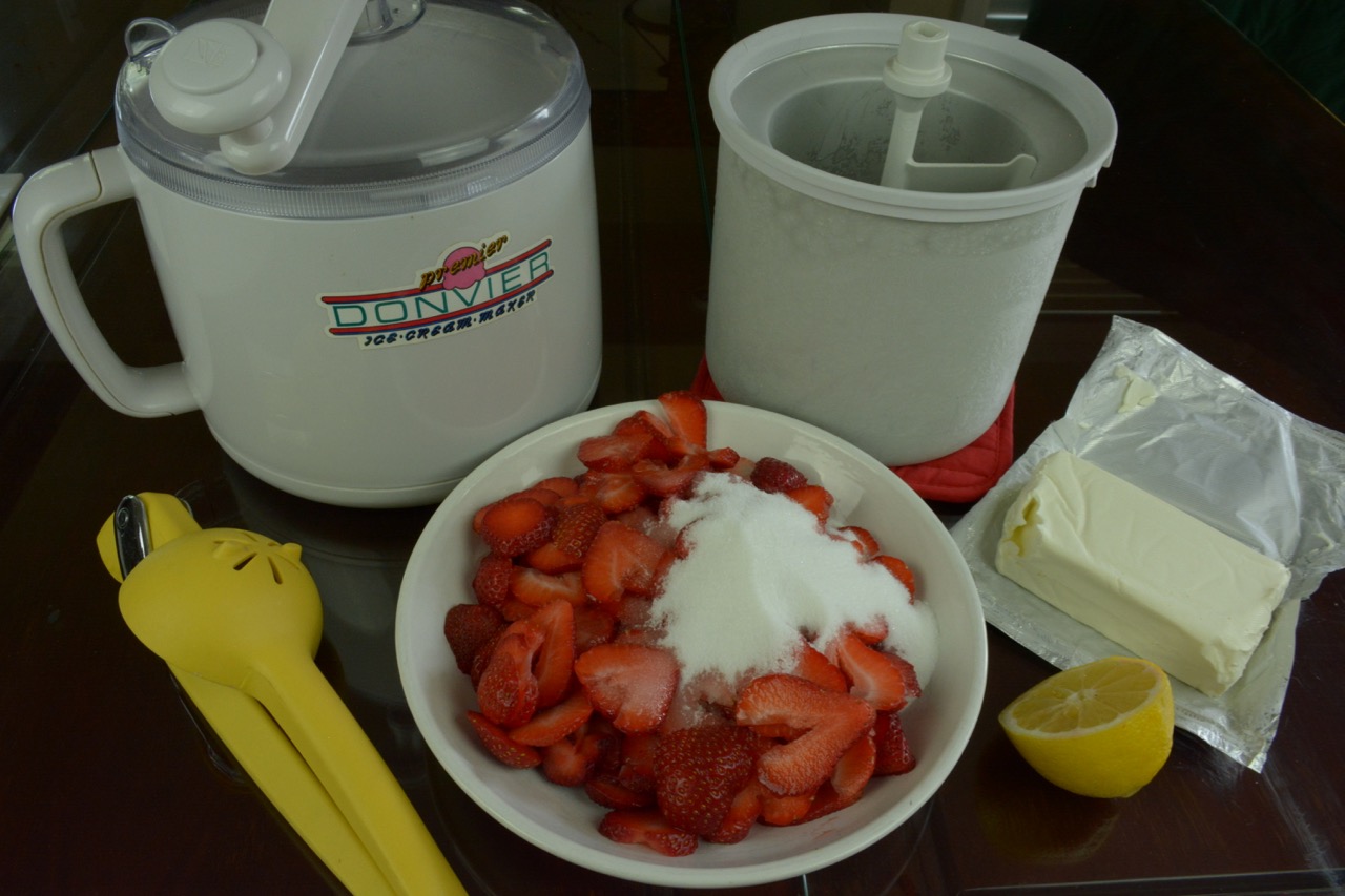 A bowl of sliced strawberries with sugar, a block of cream cheese and half a lemon on the table in front of a hand crank ice cream maker.