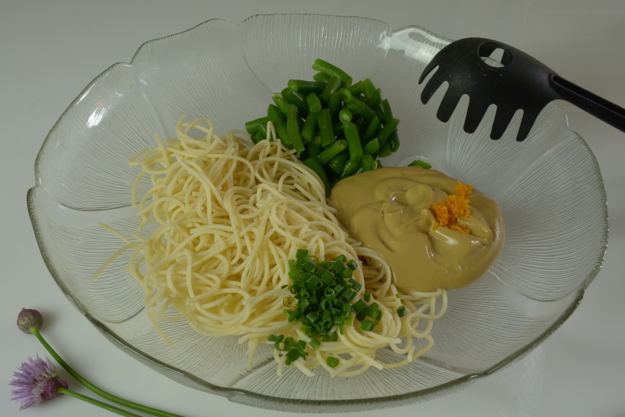 A glass bowl with cooked noodles, cooked green beans, sesame mayo, orange zest and snipped fresh chives. Beside are 2 chive stems, one with the blossom open.