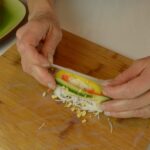 A cutting board with 2 hands starting to wrap a rice paper wrapper around a filling of noodles, peppers, cucumber and peanuts.