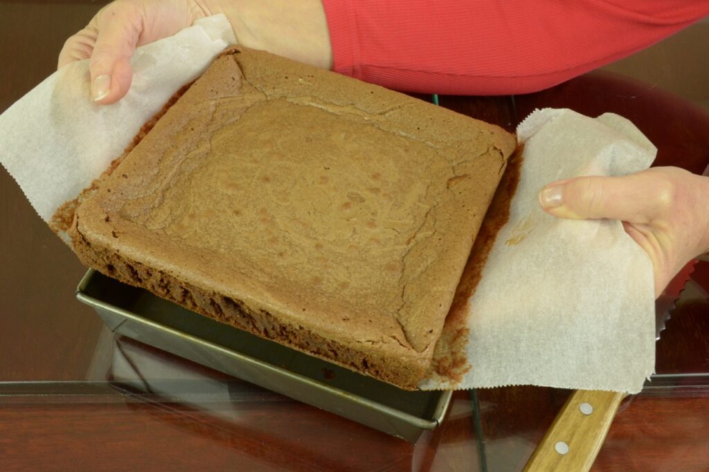 Two hands lifting a cooked brownie out of a square baking pan using the overhang of the parchment it was lined with.