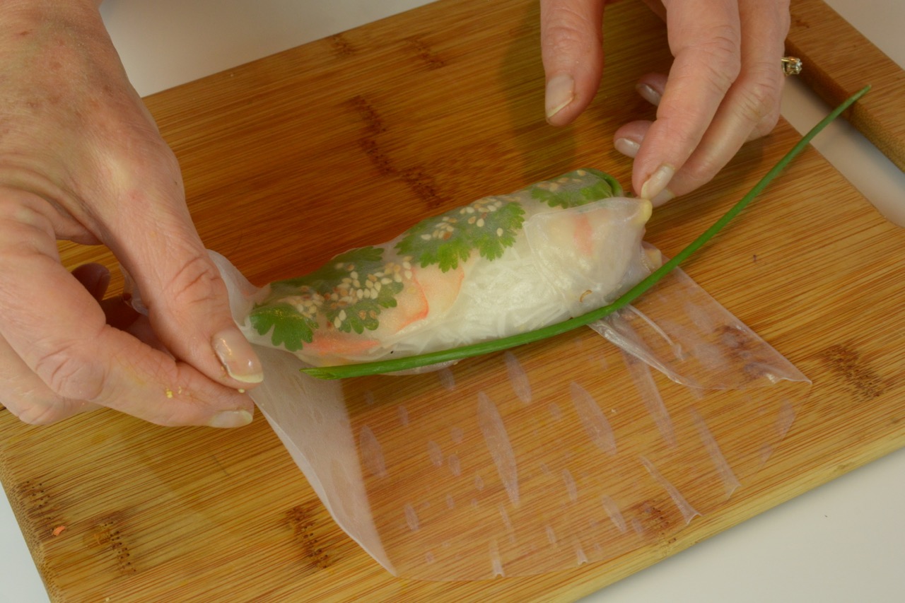 A cutting board with a salad roll showing how to add a long chive stem before the final wrap for a nice presentation.rice paper