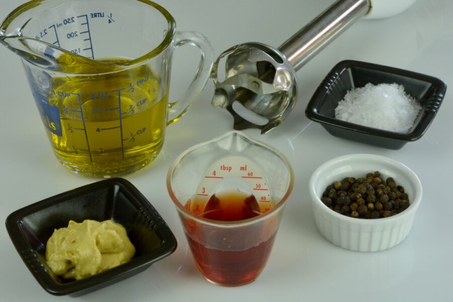 The basic ingredients to make salad dressing; oil, vinegar, mustard, salt and pepper with a hand blender beside it all.