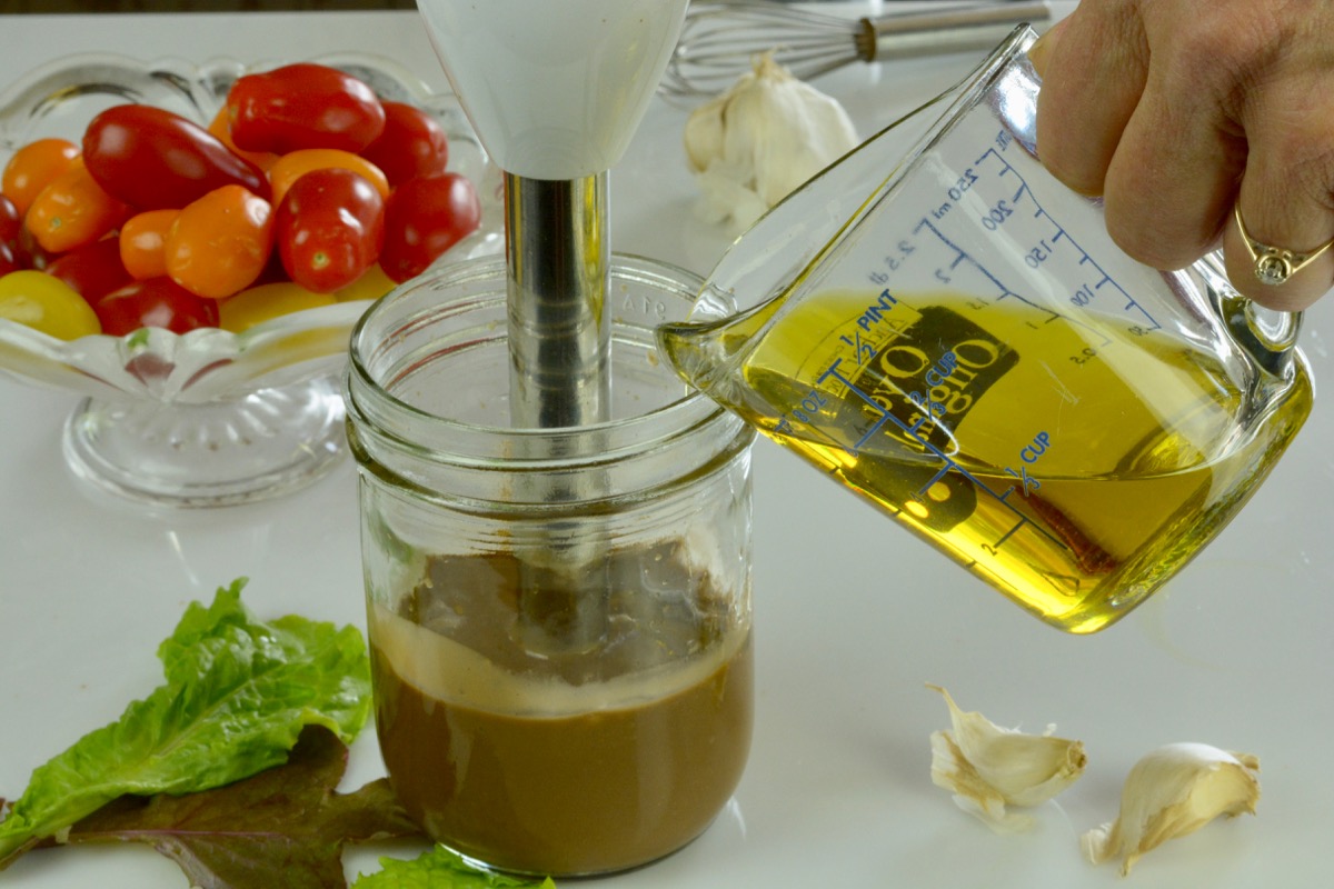 A hand blender in a Mason jar with a cup of olive oil being drizzled into it.