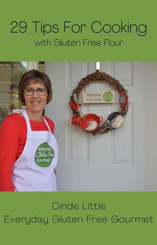 Cinde wearing her Everyday Gluten Free Gourmet apron holding the door to her house open showing a kitchen wreath that says Welcome to my Kitchen.
