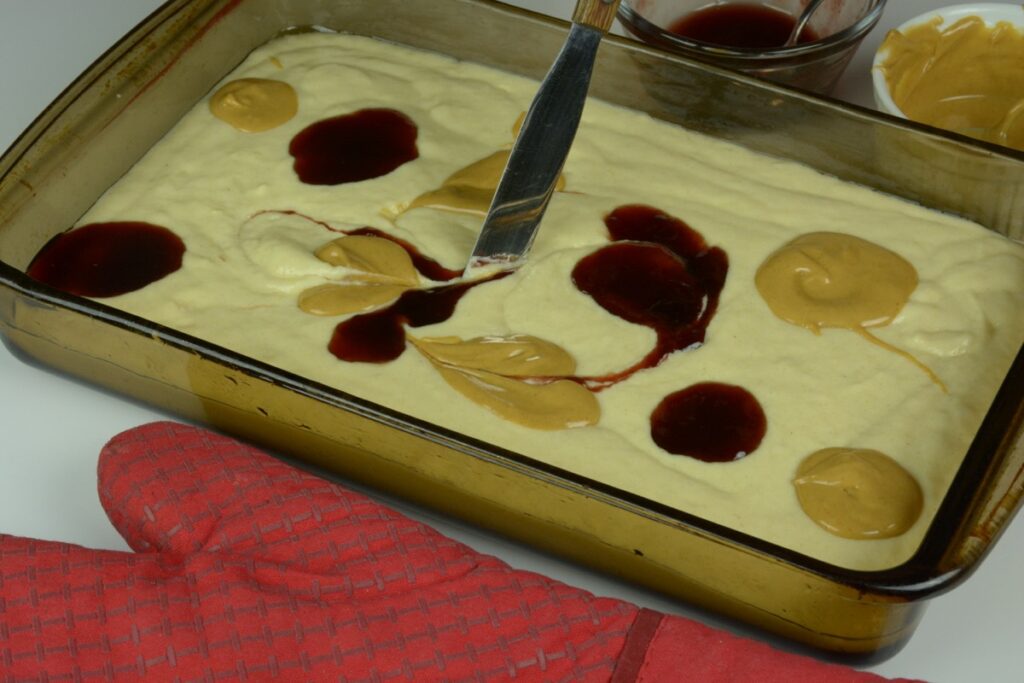 A baking pan of pancake batter topped with blobs of peanut butter and jam; with a knife starting to make swirls from them.