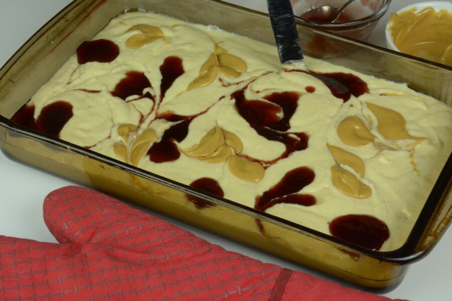 A baking pan of pancake batter topped with blobs of peanut butter and jam; with a knife starting to make swirls from them.