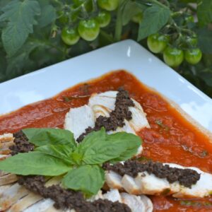 A platter of chicken slices laying on Tomato Sauce topped with Olive tapenade and fresh basil leaves.