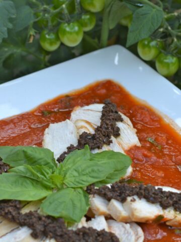A platter of chicken slices laying on Tomato Sauce topped with Olive tapenade and fresh basil leaves.